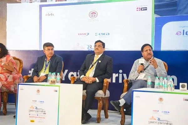 मंच पर कुर्सियों पर बैठे तीन पुरुषों में से एक माइक्रोफोन से बोलते हुए, सामने पानी की बोतलें और सम्मेलन के बोर्ड रखे हैं